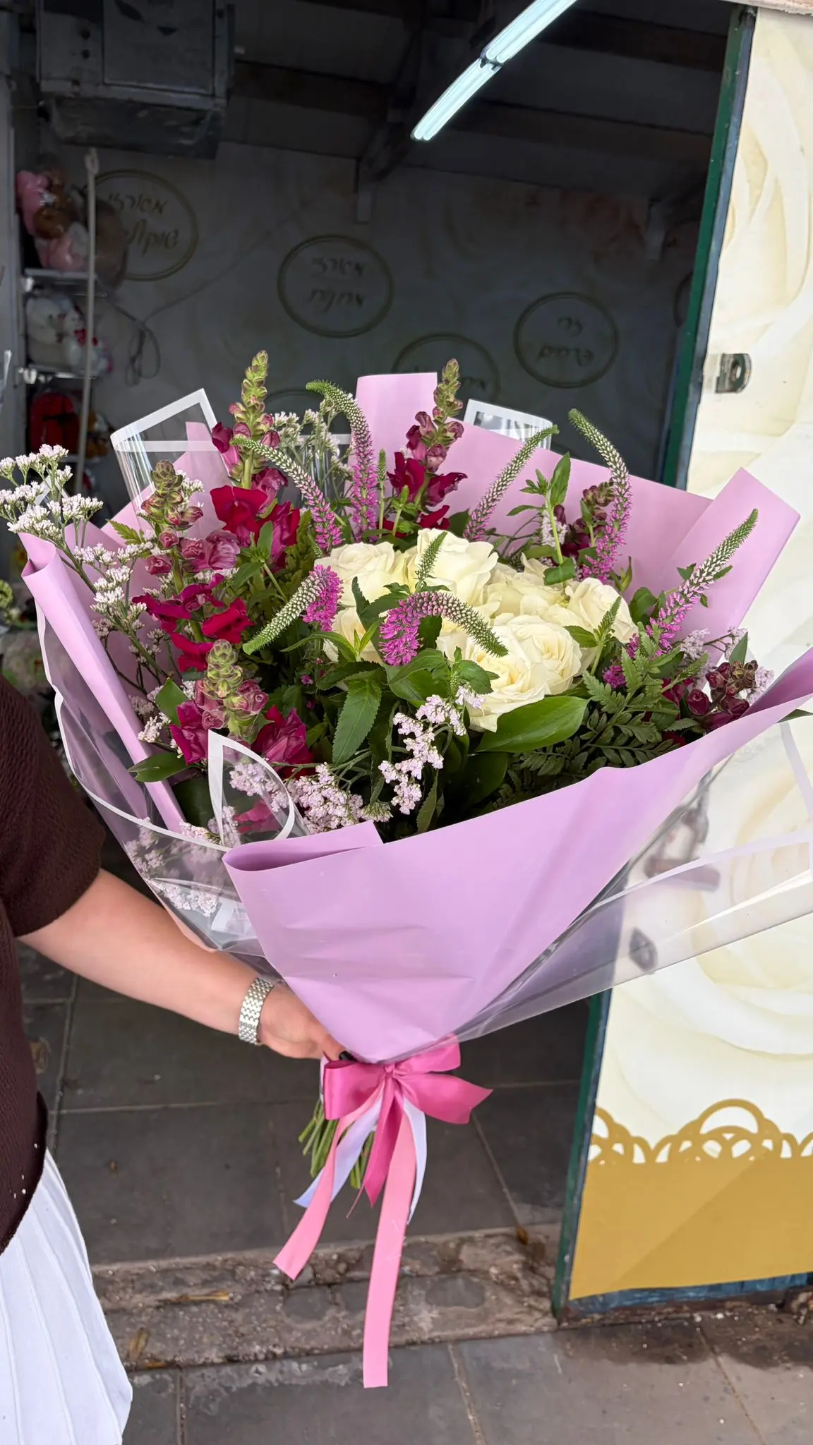 זר גדול של ורדים לבנים ופרחים ורודים וסגולים עטוף נייר ורוד עם סרט